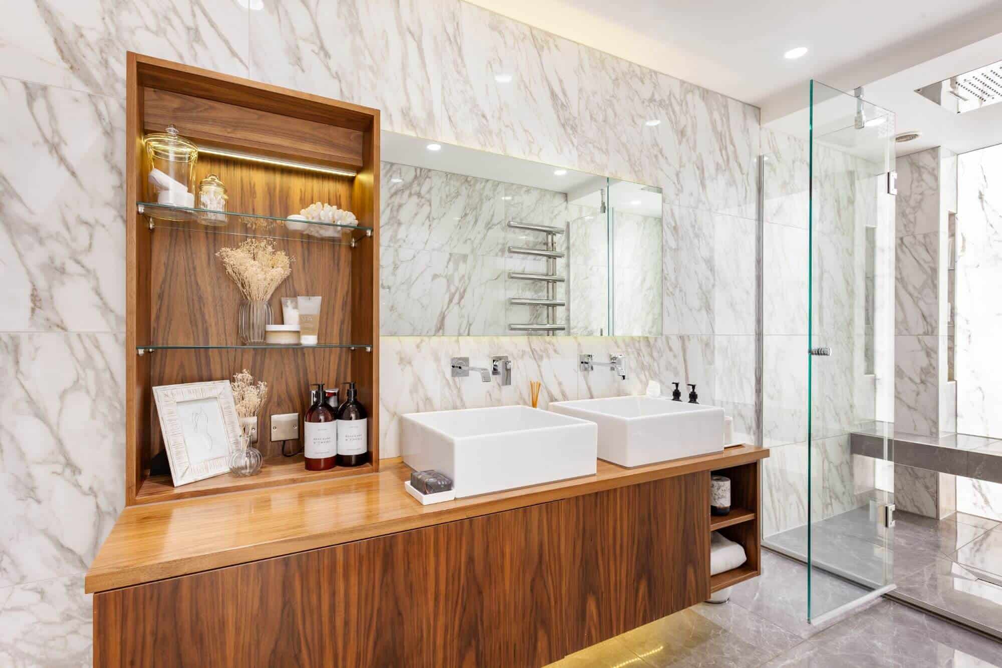 Modern bathroom with floating wall unit containing shelves, sotrage space and two wash basins. The room also contains a walk-in shower with glass doors and marbled wall and floor tiles