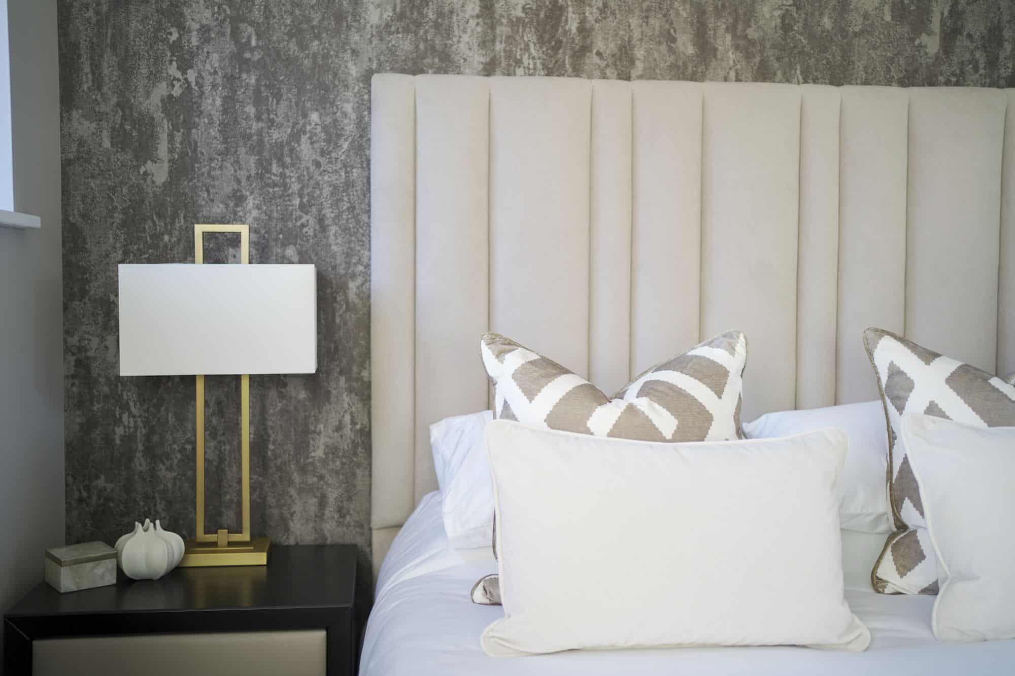 Left side of a double bed with neutral colour covers and cushions, with a dark grey bedside table next to it, featuring a cream-coloured drawer and a modern gold-coloured rectangular lamp with white shade
