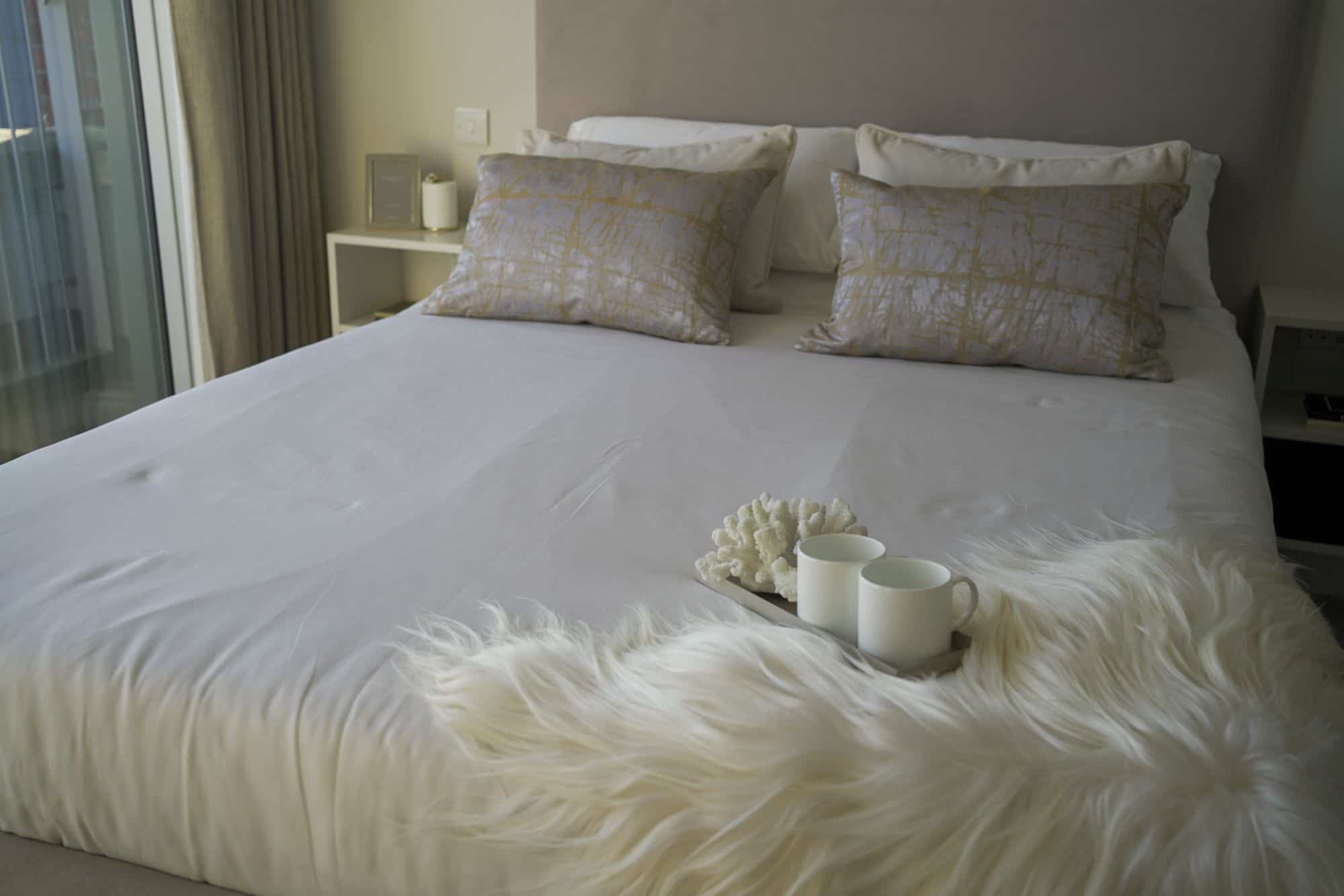 Double bed in a master bedroom with neutral colour covers and cushions, with a coffee mug tray and white fur-style rug at the bottom of the bed