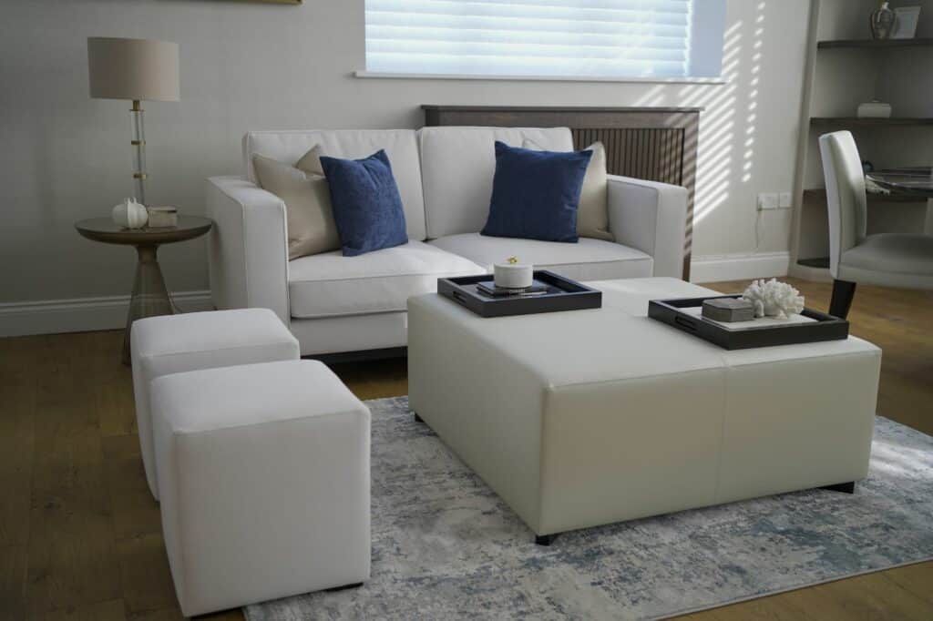 A modern sitting room in muted colours with a modern sofa, glass-based side table, upholstered coffee table and two footstalls